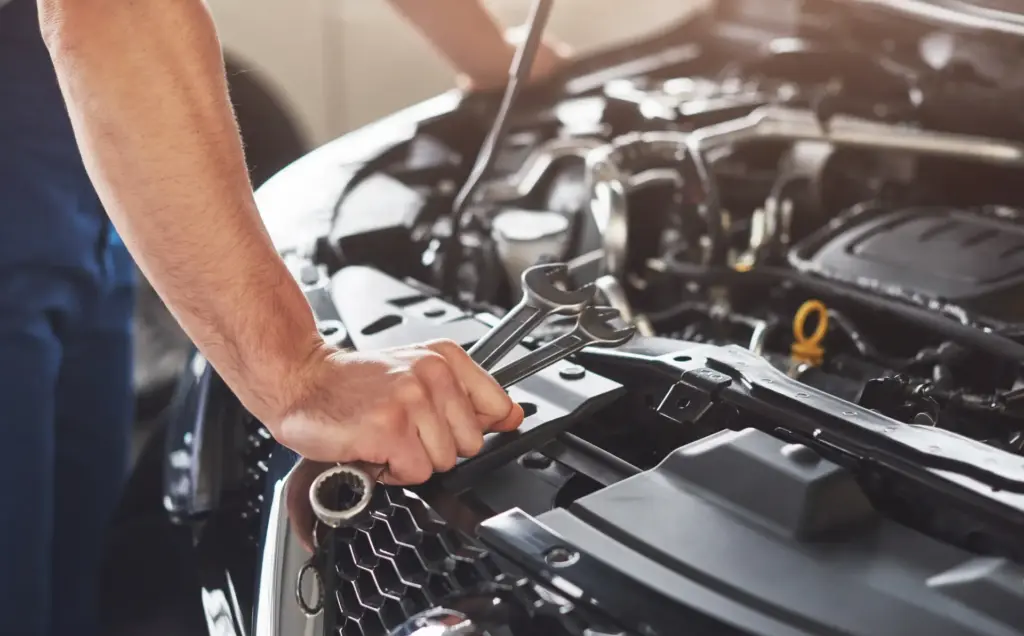 Mechaniker hält Schraubenschlüssel, während er in einer Werkstatt eine Autoinspektion an einem Automotor bei geöffneter Motorhaube durchführt.
